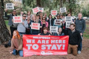 a group of tenants and allies of LATU Hollywood gather in a park and hold up signs that say "RENT STRIKE" and a red-and-white banner that reads "We are here to stay!"