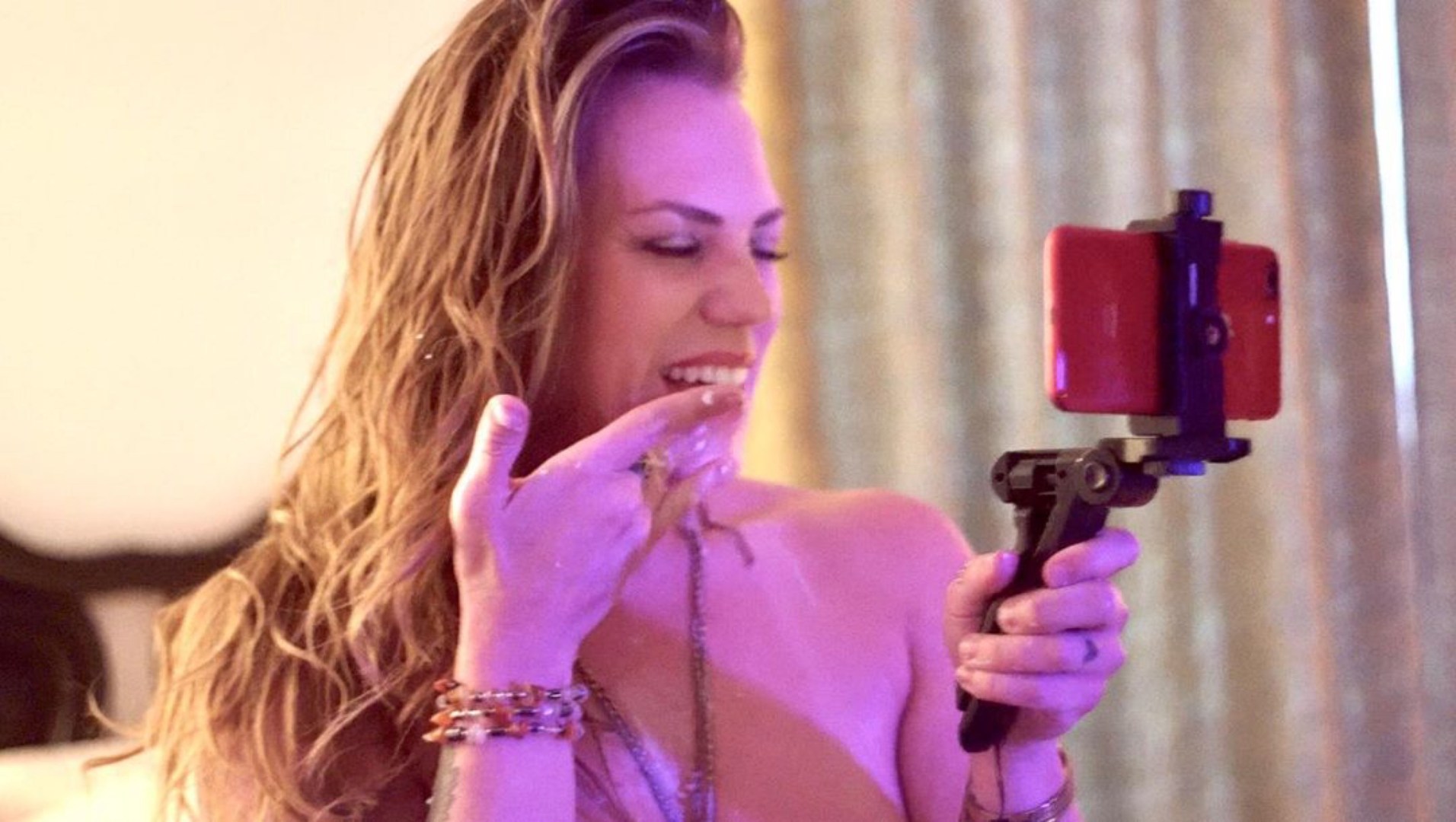 A strawberry blond woman holds a finger to her mouth and smiles for the camera on her phone she is holding with a tripod