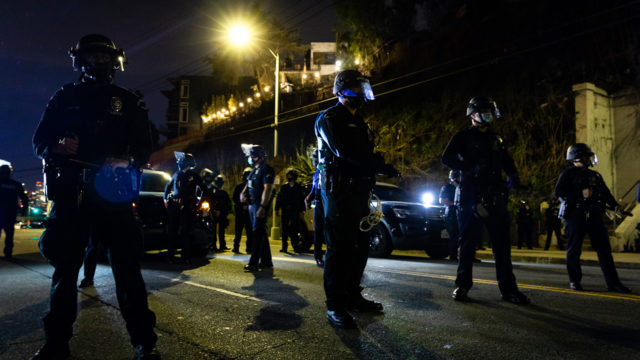 A skirmish line of LAPD officers in riot gear at night blocking the street towards Echo Park Lake