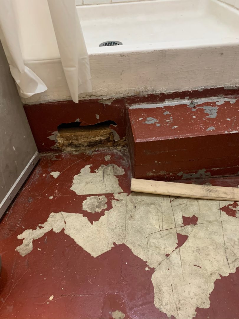 A dirty bathroom floor next to a shower in a Healthy Housing Foundation facility. The red tile in the floor is chipped and dirty and there is a large hole between the shower and the floor. 