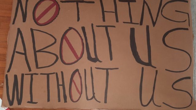 A sign made from a piece of cardboard. It reads "Nothing About Us Without us" in black permanent marker:.
