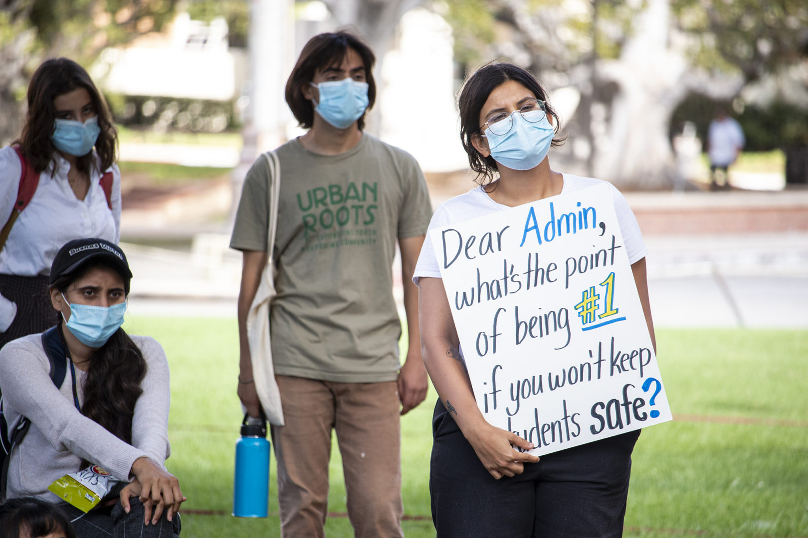 Students wearing face masks take part in the protest.  One with glasses is holding a sign saying 