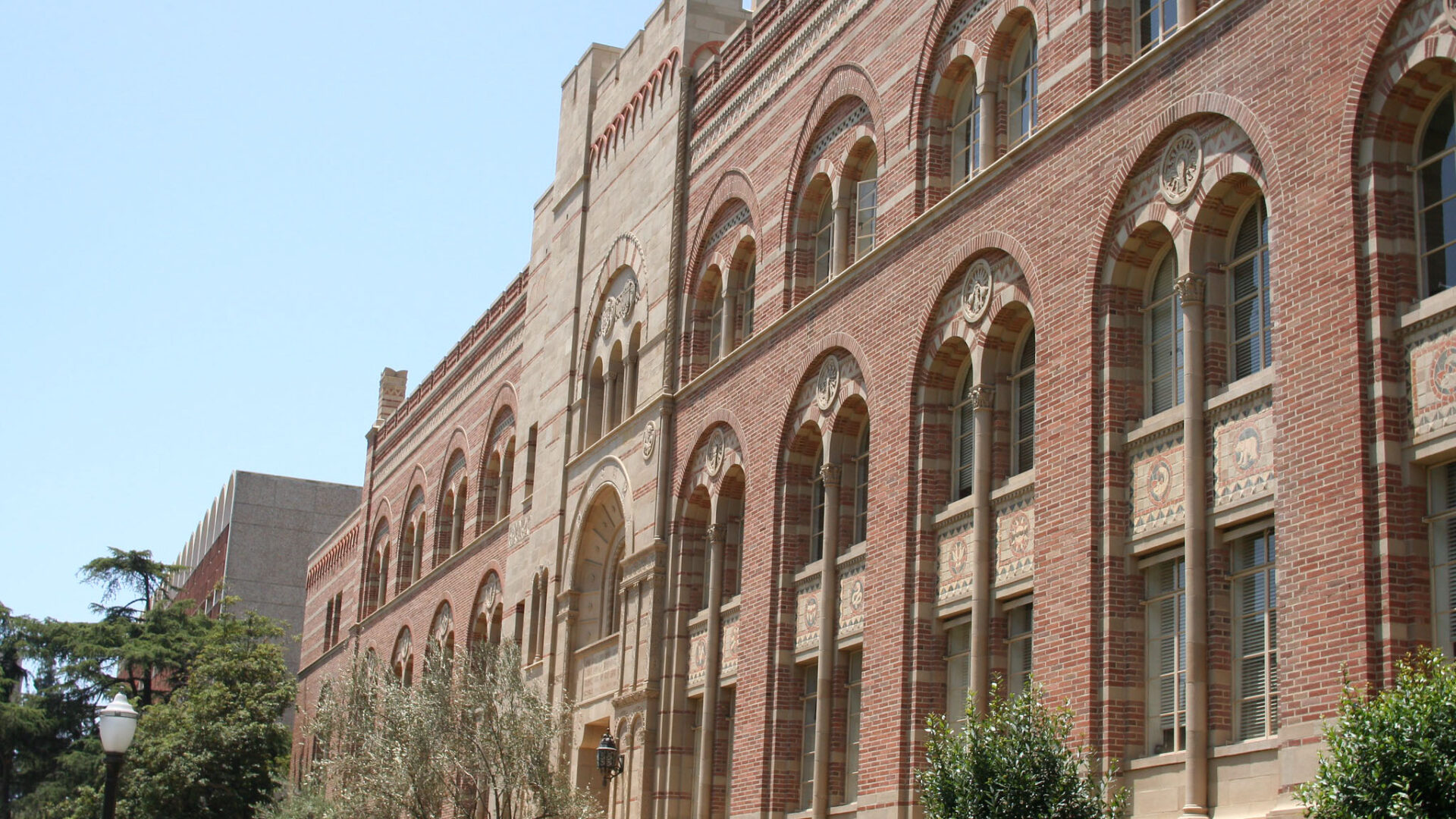 The brick exterior of a University of California, Los Angeles building. Trees line the lower wall. 