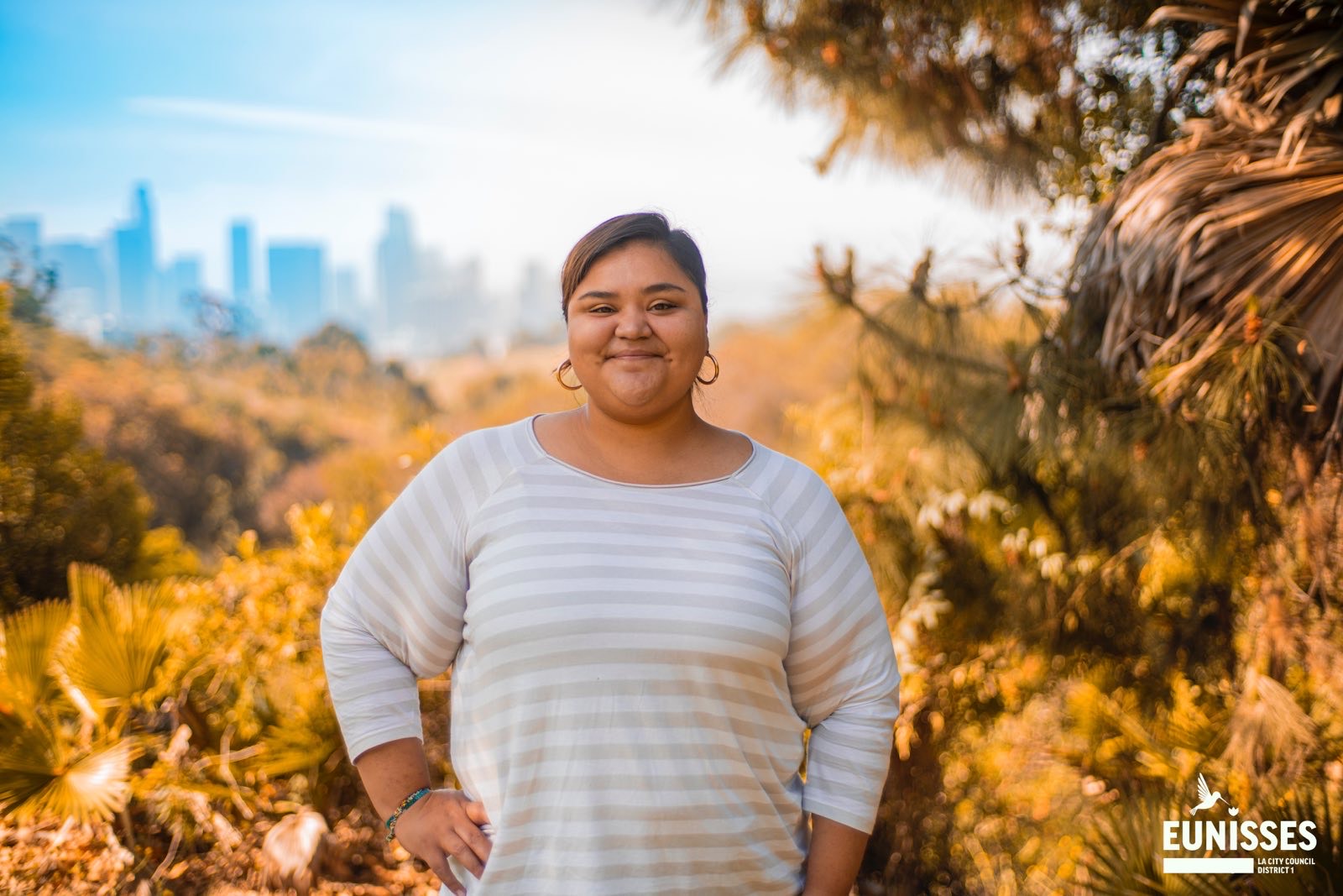 Council District One Candidate Eunisses Hernandez is standing in a softly lit chaparral with trees and bushes smiling, with downtown Los Angeles in the background and a campaign logo on the bottom right.