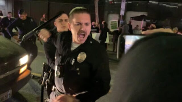 an LAPD officer yells as he prepares to swing his baton at a protester holding a camera phone. Several other LAPD officers behind him look on.