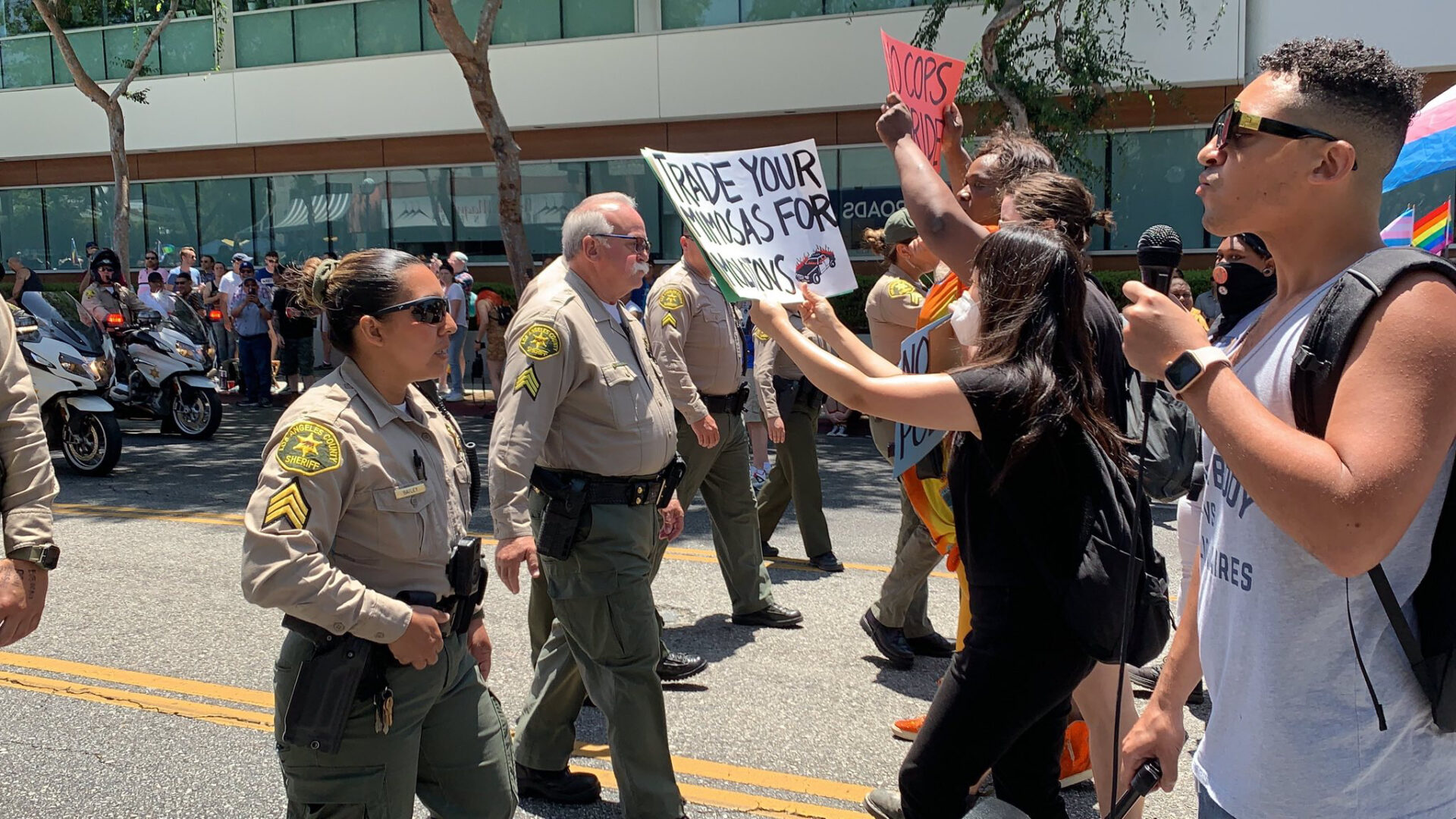 Protesters are being back down by a group of Los Angeles Sheriff's Deputies at LA Pride. One protester is holding a microphone and two are holding signs. One sign says "No cops at Pride" and the other says "Trade your mimosas for molotovs."