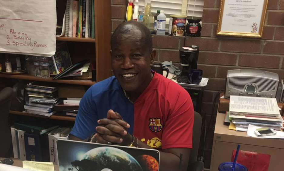 Principal Gilberto Samuel sits at a desk in a cluttered room. His hands are clasped.