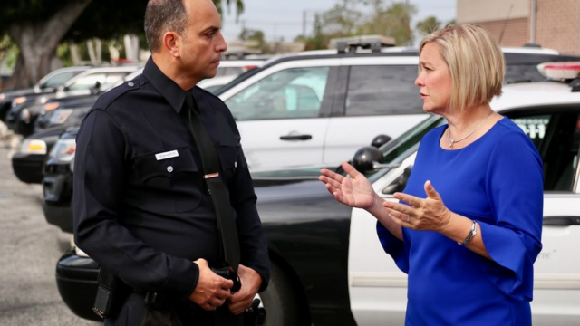 Traci Park, wearing a blue dress, speaks with a policeman in uniform in front of a line of parked police cruisers