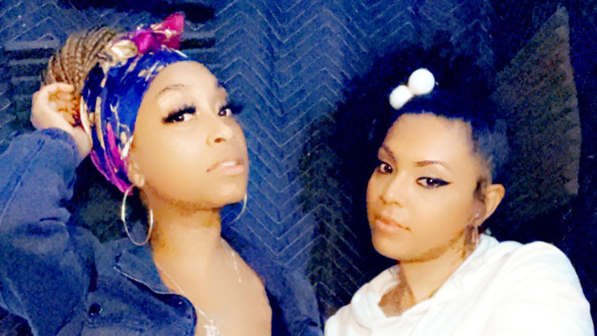Mz Free, wearing a blue and purple head wrap, poses with YaH-Ra, who is wearing a white shirt. The two of them are in a recording studio.