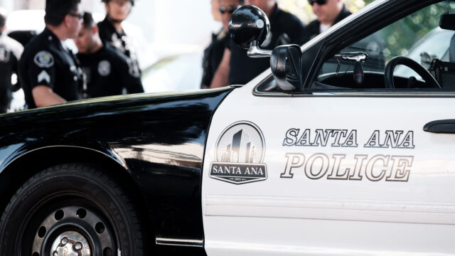 The front half of a black and white Santa Ana police car in the foreground with individuals wearing police uniforms and sunglasses in the background.
