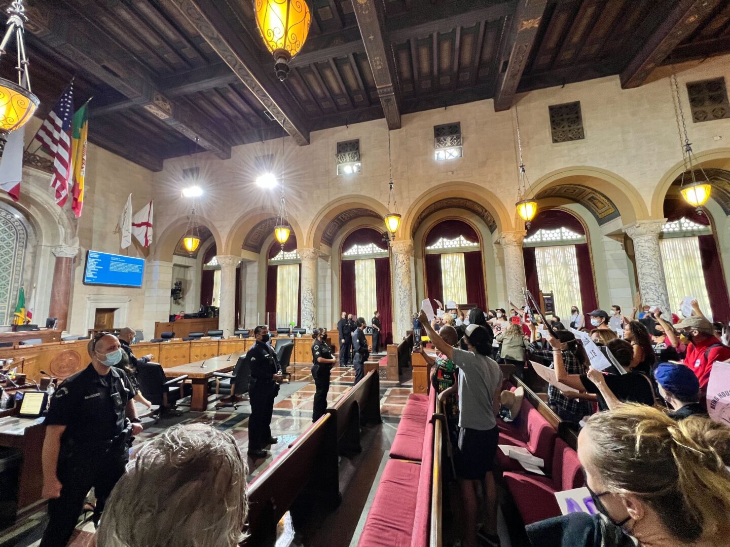 LA City Council meeting with audience holding up signs and police officers facing them from the front of the room.