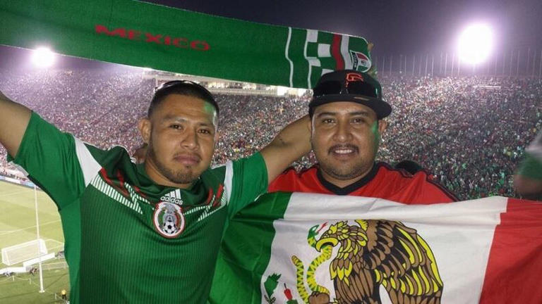 Two men stand in front of a soccer field in a crowded stadium. The man on the left wears a green Mexico soccer jersey and holds a scarf above his head while the man on the right holds the flag of Mexico.