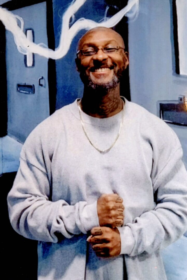 Ajamu wearing glasses, a gold chain, and a light gray sweater and smiling into the camera.