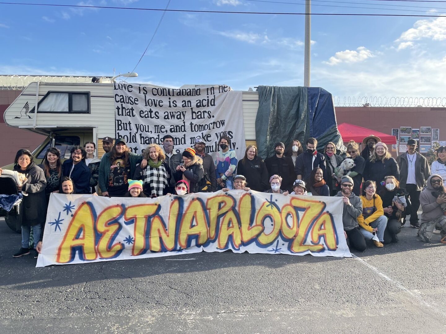 Around 30 people stand in front of an RV in the daytime with a sign that reads "Aetnapalooza."