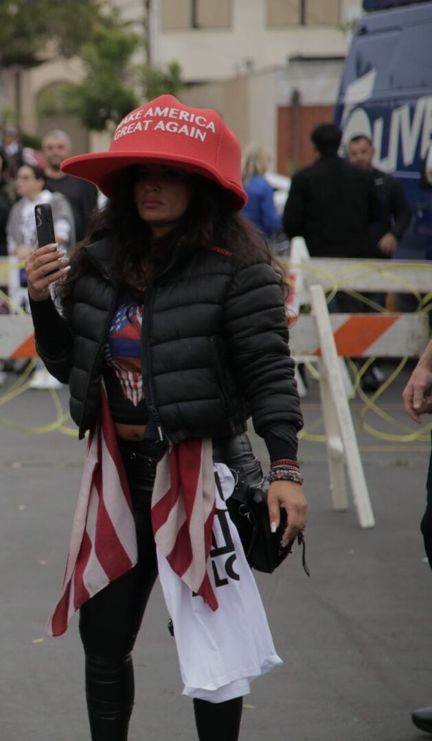 A woman wears an oversized MAGA hat and wields t-shirts in her belts, for some reason. 