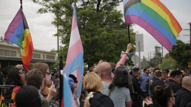 LGBTQ+ advocates and counterprotesters hoist large Progress Pride flags and the transgender Pride flag above a large group flanked by police officers.