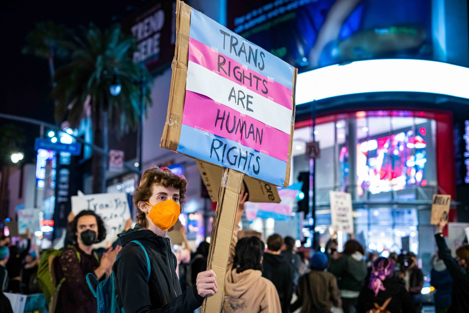 Activist with an orange N95 mask holding a sign with the trans flag saying "Trans Rights are Human Rights"