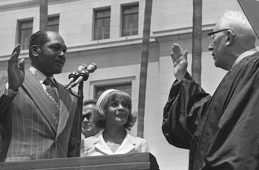 A black and white photograph of Tom Bradley standing at a podium and getting sworn in as the mayor of Los Angeles.