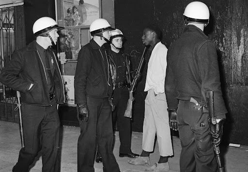 A black and white photograph of four armed policemen surrounding a Black resident. One policeman is holding up their gun towards the resident. The photo was taken in 1966