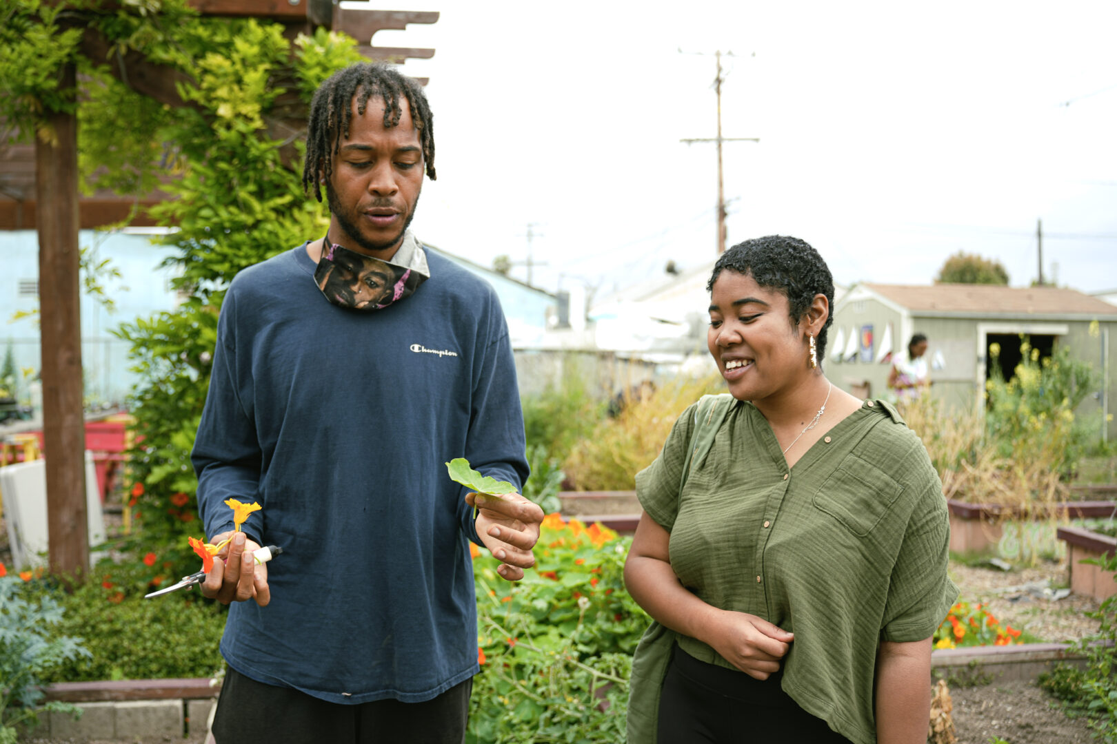 Two people at a garden speaking while holding plants. The man on the left is holding a yellow flower and a green plant. 