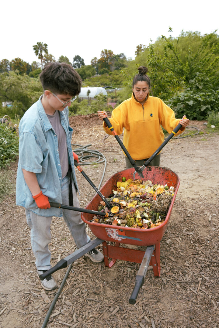 Two individuals are pictured working at a composting center. They have giant tools in their hands and are slicing up waste in a red wheelbarrow.