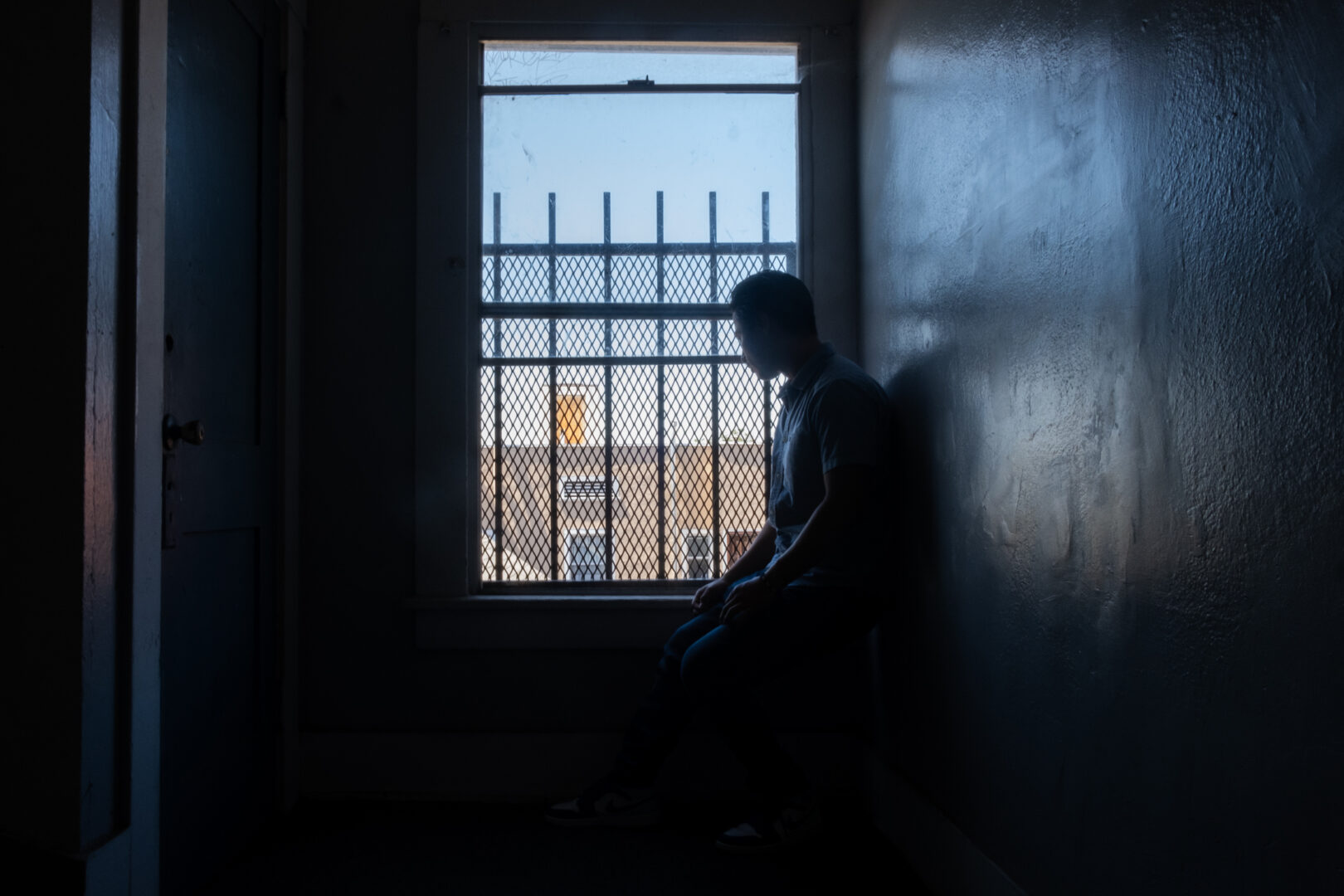 In the shadows, Kent Mendoza looks out the window located in a hallways of the apartment building he grew up in. July 2023. Credit: Rodrigo Magaña | Knock LA