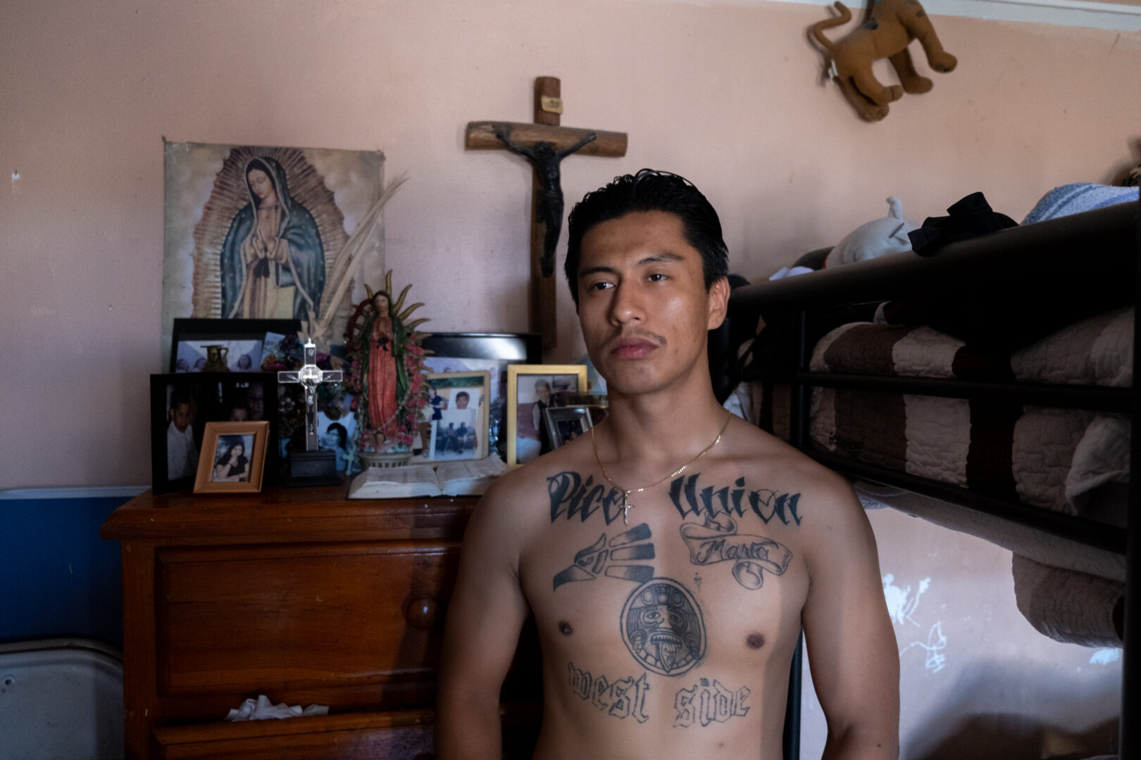 Kent Mendoza in front of his mother’s altar showing his tattoos that represent his community, a place he is proud of being from. July 2023. Credit: Rodrigo Magaña | Knock LA
