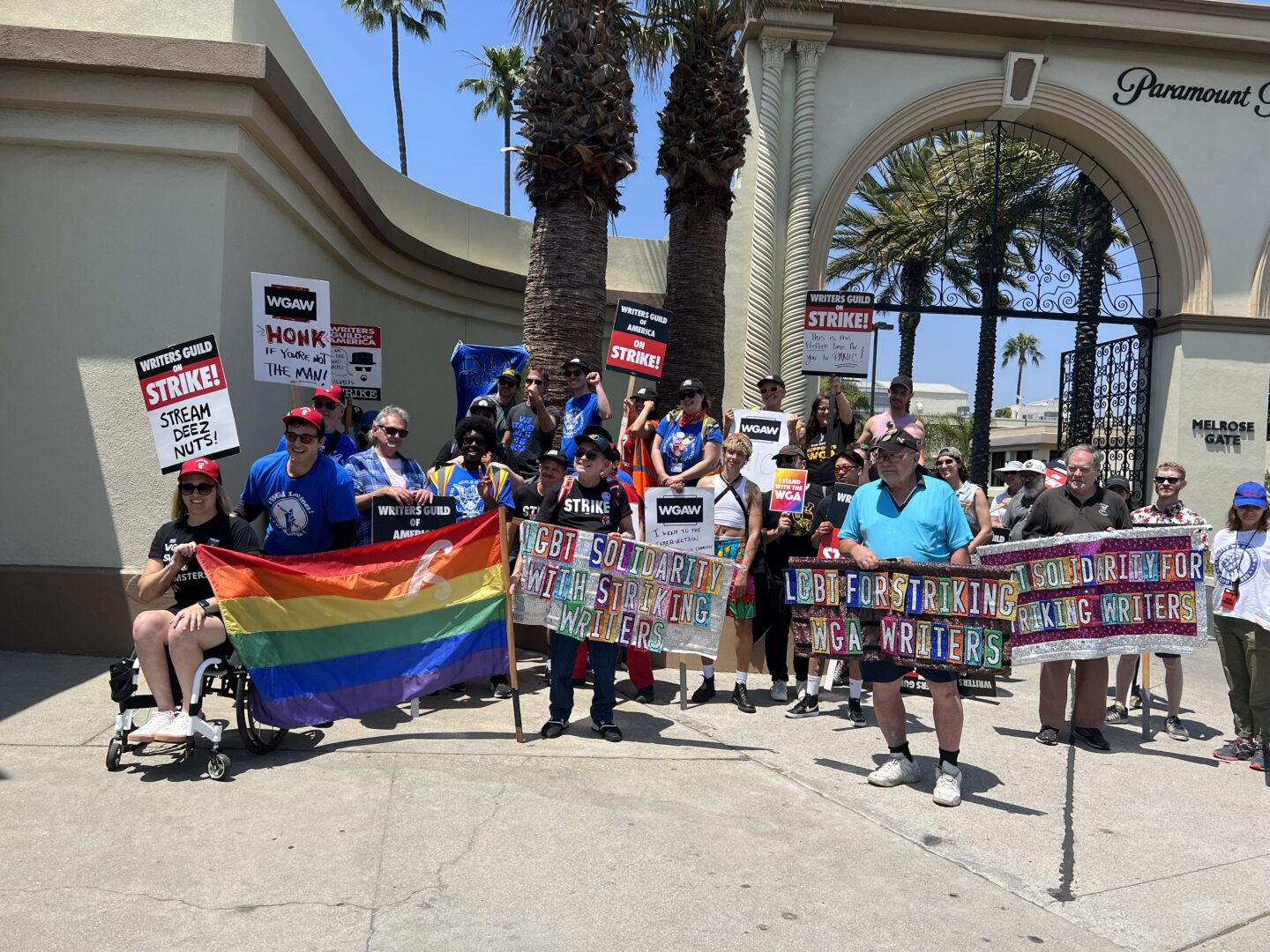 People striking in front of Paramount Studios carrying signs that say "LGBT For Striking WGA Workers" and "WGA" signs