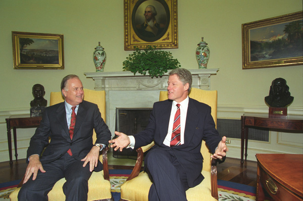 A photograph of Richard Rioran and Bill Clinton, wearing black suits and red ties, seated next to each other in an office.