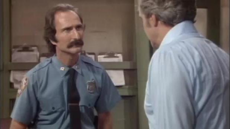 Officer Zatelli, a white man with brown hair and a receding hairline sporting a bushy mustache, is wearing a blue policeman's uniform with a black tie. He is standing in a police station, and is looking at another man with his back to the camera. The other man is white and wearing a white long sleeved shirt and has light colored hair. 