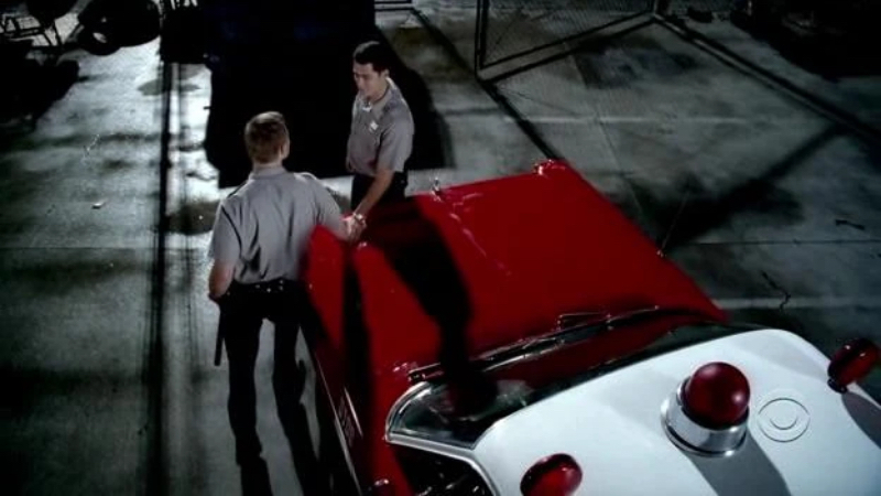 Two white men in gray and black police uniforms hold hands over a red, old-fashioned abulance parked in the street at night. 