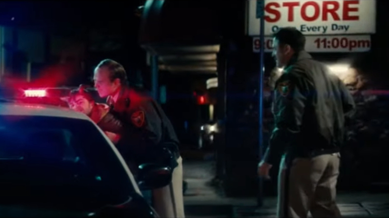 Two white police officers in uniform on a street at night. One on the left of the screen is leaning his body against a person he he pushing into a patrol car. 