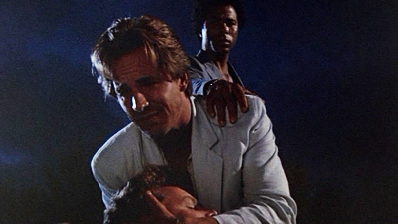 A white man with brown shaggy hair wearing a gray suit cries as he cradles another man in his arms. Behind him, a Black man in a black suit with a short afro puts his hand on the crying man's shoulder. 
