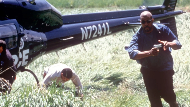 A helicopter dominates the frame. Underneath the tail, a man in a long sleeved light colored shirt is hunched over. A Black man pointing a gun to the right of the frame runs forward towards the camera. He has a bald head and is wearing sunglasses, a navy windbreaker with a similarly colored shirt underneath, and black plants. 