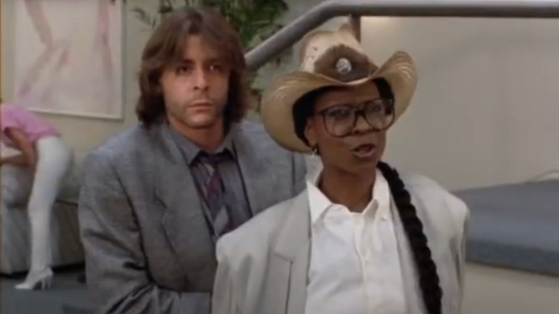 A Black woman in a straw cowboy hat with her hair in a long braid grimaces at the camera. She is wearing large brown framed glasses, a white collared shirt and gray blazer. Behind her is a white man with long brown in a gray suit.