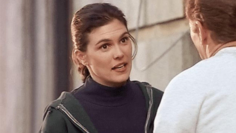 Abby Sullivan, a white woman with brown hair, stands in front of another person talking to them. They are both shown from the shoulders up. Her hair is pulled back into a low ponytail, and she is wearing a black turtleneck with a green hooded sweatshirt that is slightly unzipped. 