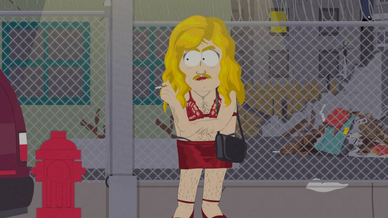 Sergeant Yates, a white cartoon man, is in drag standing on the street in front of a chain link fence. He is wearing a blonde wig, red lace top and red low slund skirt with his thong showing. He has stubble visible on his leg, has a blonde mustache and carries a black purse. He is wearing red strappy heels. 