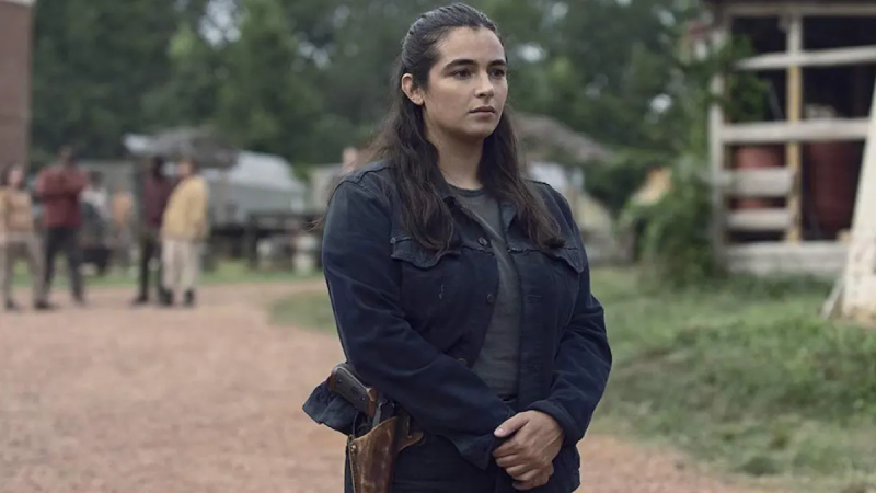 Tara Chambler, a fair skinned woman with brown hair falling just past her shoulders, stands in a rural street on a dirt road. She is wearing a dark jacket with a grey undershirt. She carries a gun on her hip in a brown leather carrier. 